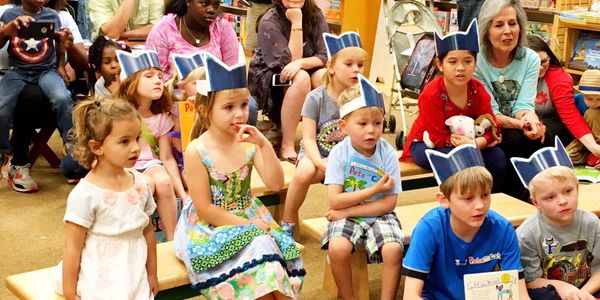 Young children and adult volunteers are sitting in a group while a book is read to the group.