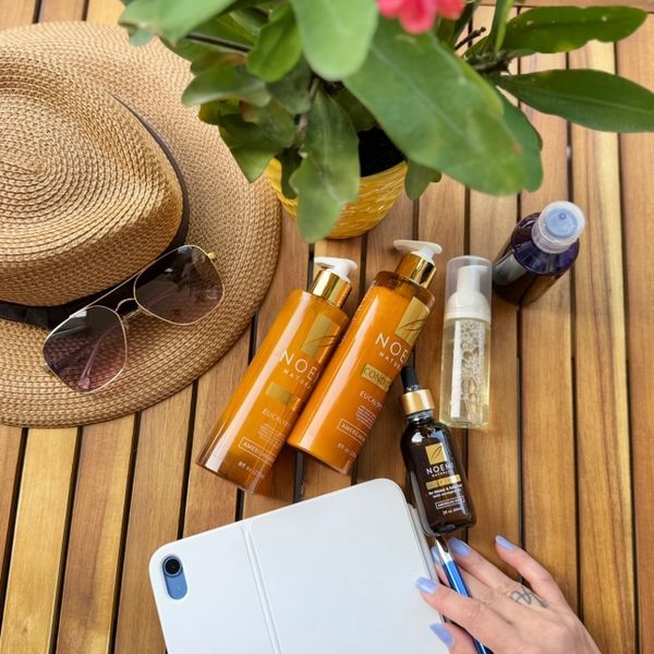 Sun hat with sunglasses, skincare products, and a hand holding a tablet on a wooden table.