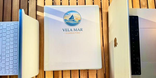 A binder with Vela Mar Consulting logo between two laptops on a wooden table.