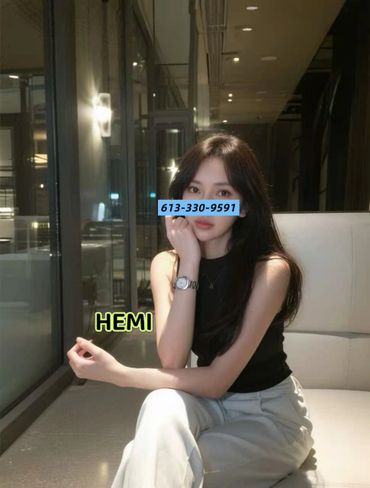A woman with long dark hair sitting indoors near a window, wearing a black sleeveless top and white pants.