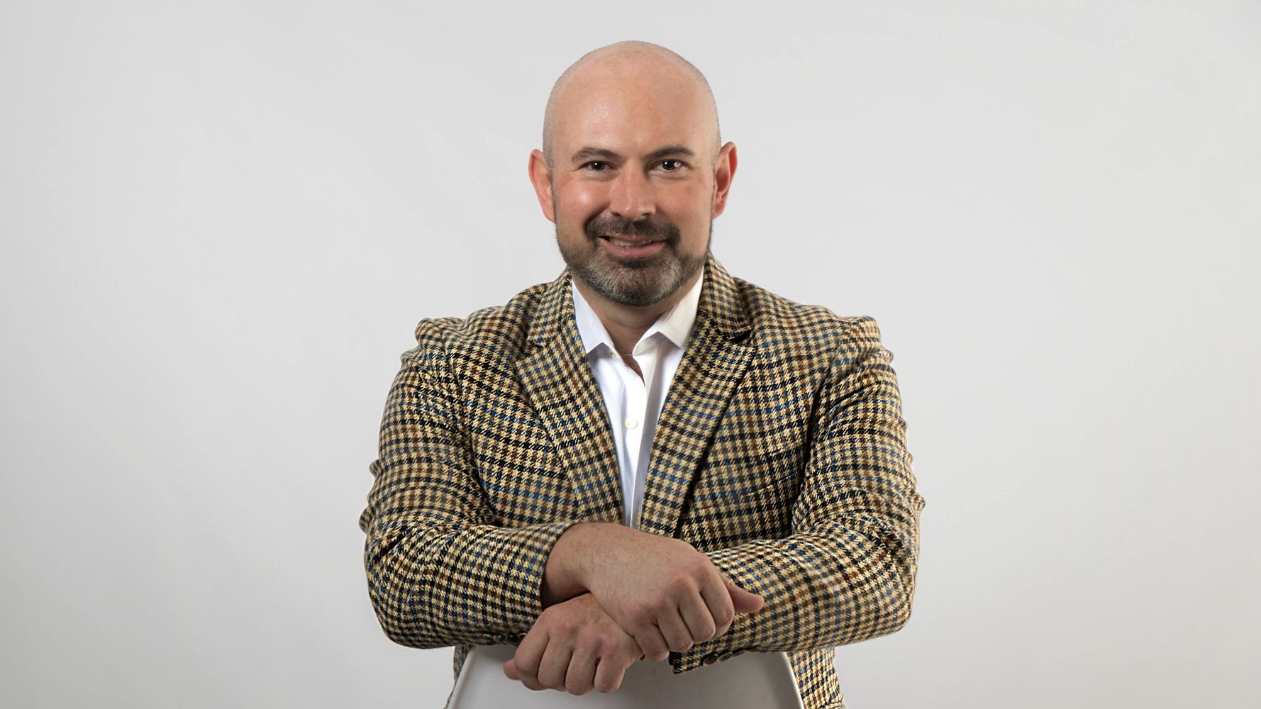 Smiling bald man in a checkered blazer sitting on a chair against a plain background.