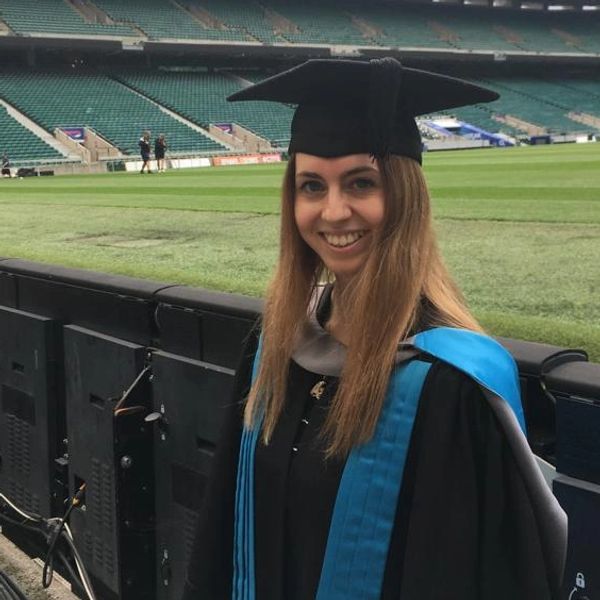 A smiling graduate in cap and gown at an empty stadium field.