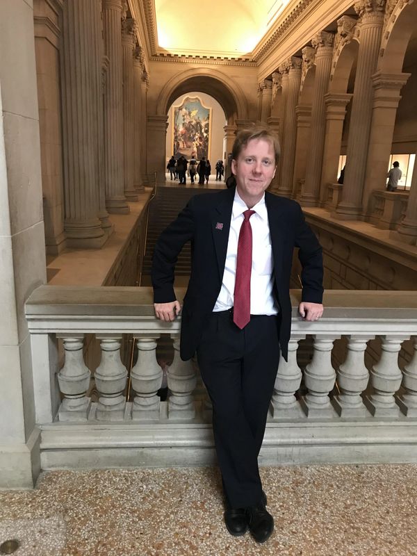 Man in a suit leaning on a stone railing inside a grand, classical building.