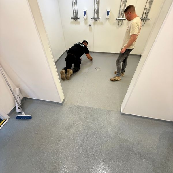 Two men working on a clean, grey floor in a white-walled room with shower fixtures.