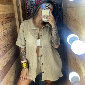 Person taking a mirror selfie wearing a beige oversized shirt and shorts set with tattoos on arms.