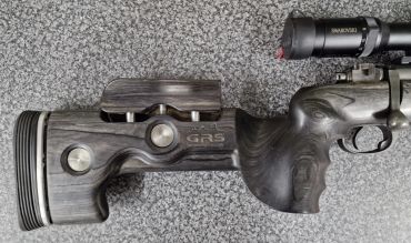 Close-up of a rifle stock with a mounted scope on a textured surface.