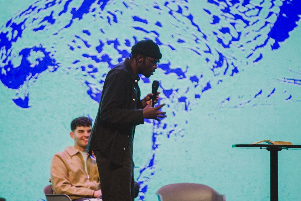 A man passionately speaking into a microphone on stage with a large eagle graphic behind him.