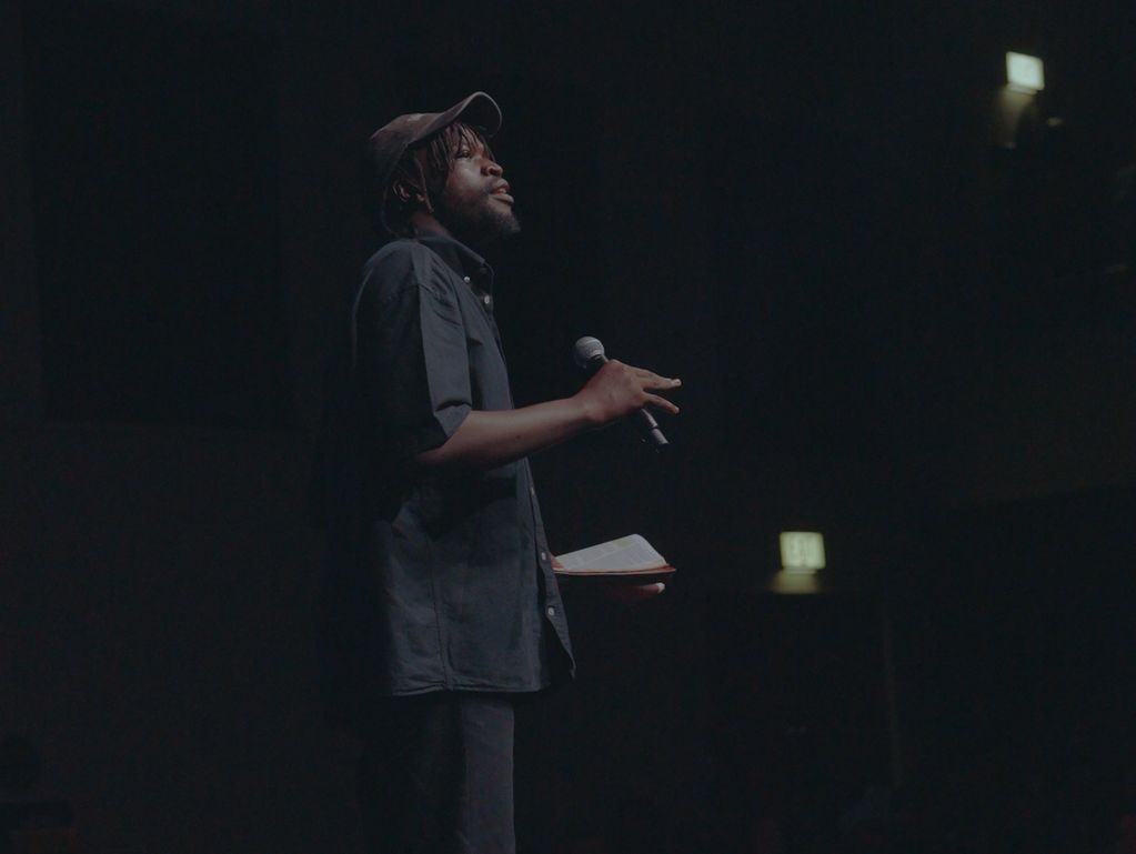 Man with a microphone and book speaks on a dark stage.