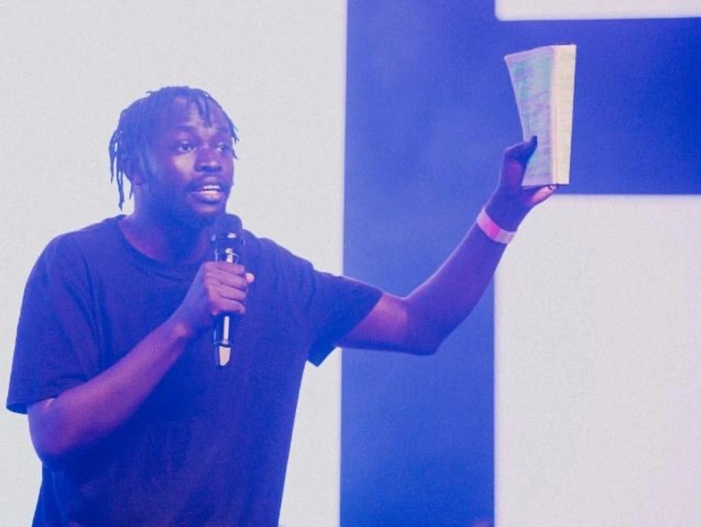 Man speaking on stage, holding a book and microphone.