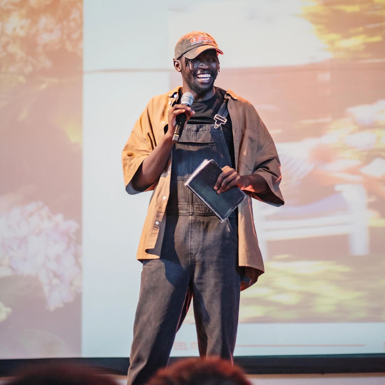 A man smiling while speaking on stage holding a microphone and a notebook.