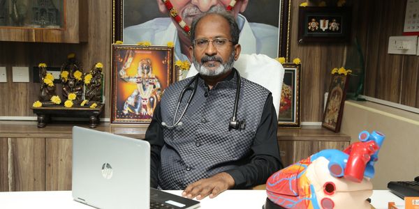 A doctor is seated at a desk in a modern hospital office, working on a laptop with a heart model placed nearby, symbolizing cardiology expertise. The hospital chairman is also present, seated across the desk, observing or discussing patient care plans. The office is well-lit and professional, reflecting a trustworthy and advanced medical environment.