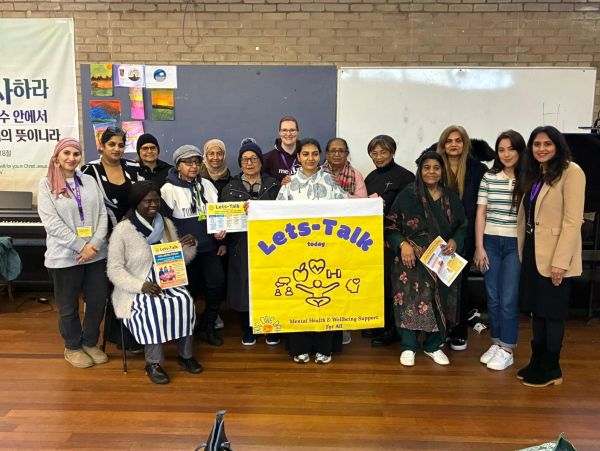 A diverse group holding a 'Lets-Talk' mental health support banner indoors.