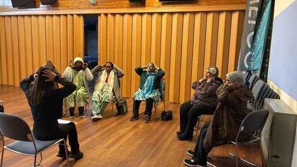 Group of elderly women doing seated exercises with instructor in a community hall.