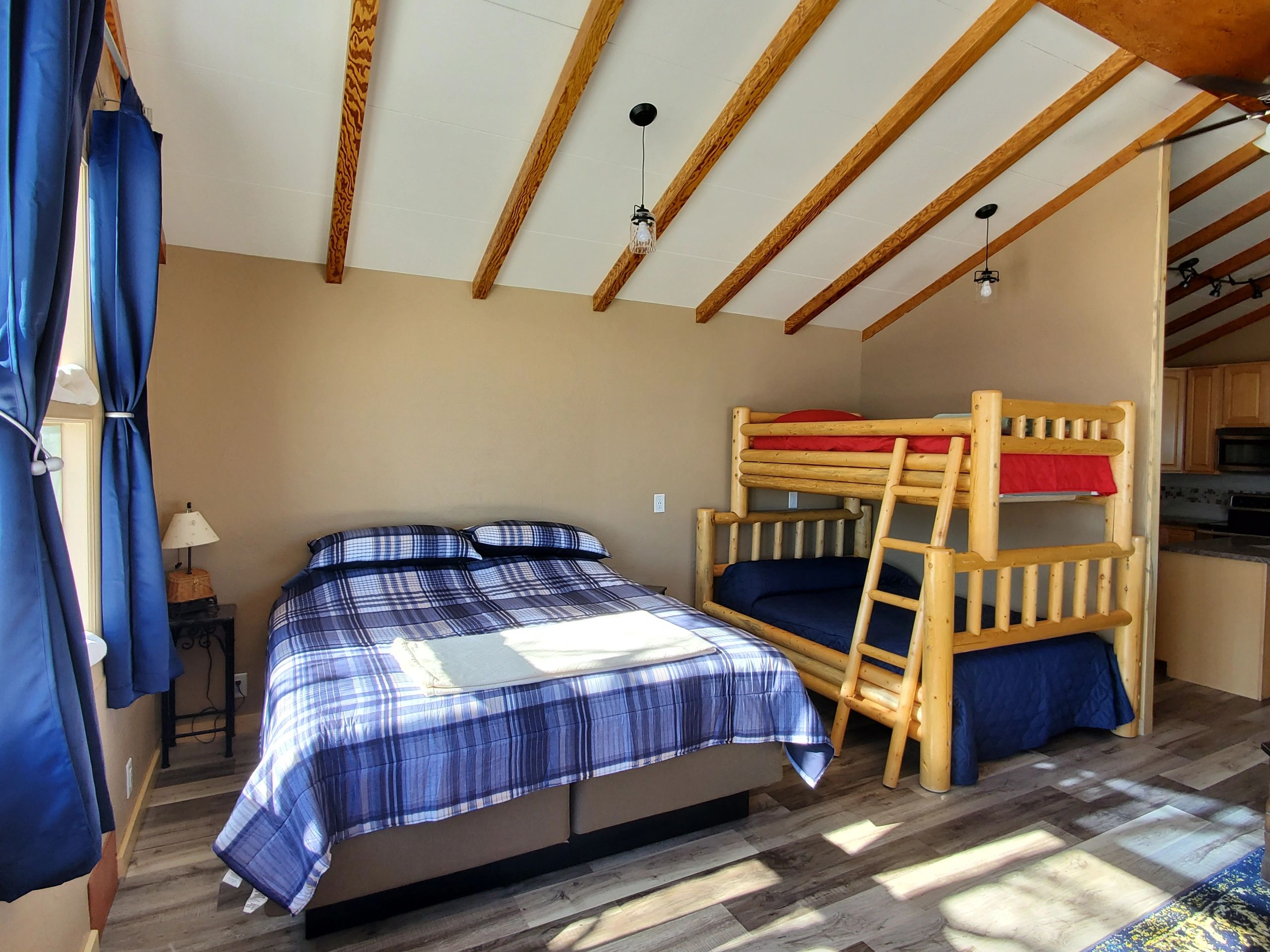 A wide view of a room with a double bed and bunk bed, vaulted ceiling with wood beams, and a kitchenette.
