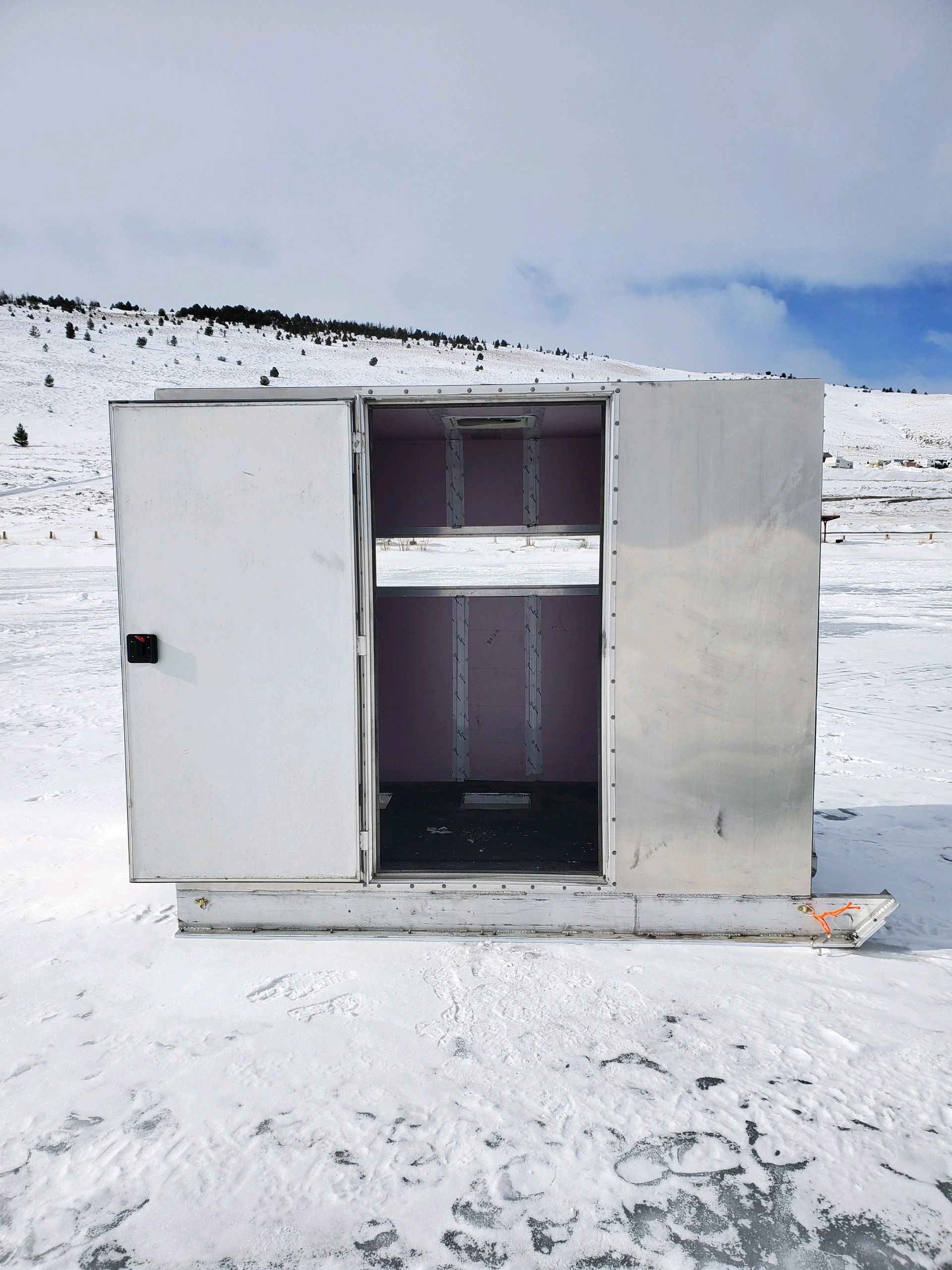 a small metal ice fishing hut with an open door sits in a snowy landscape