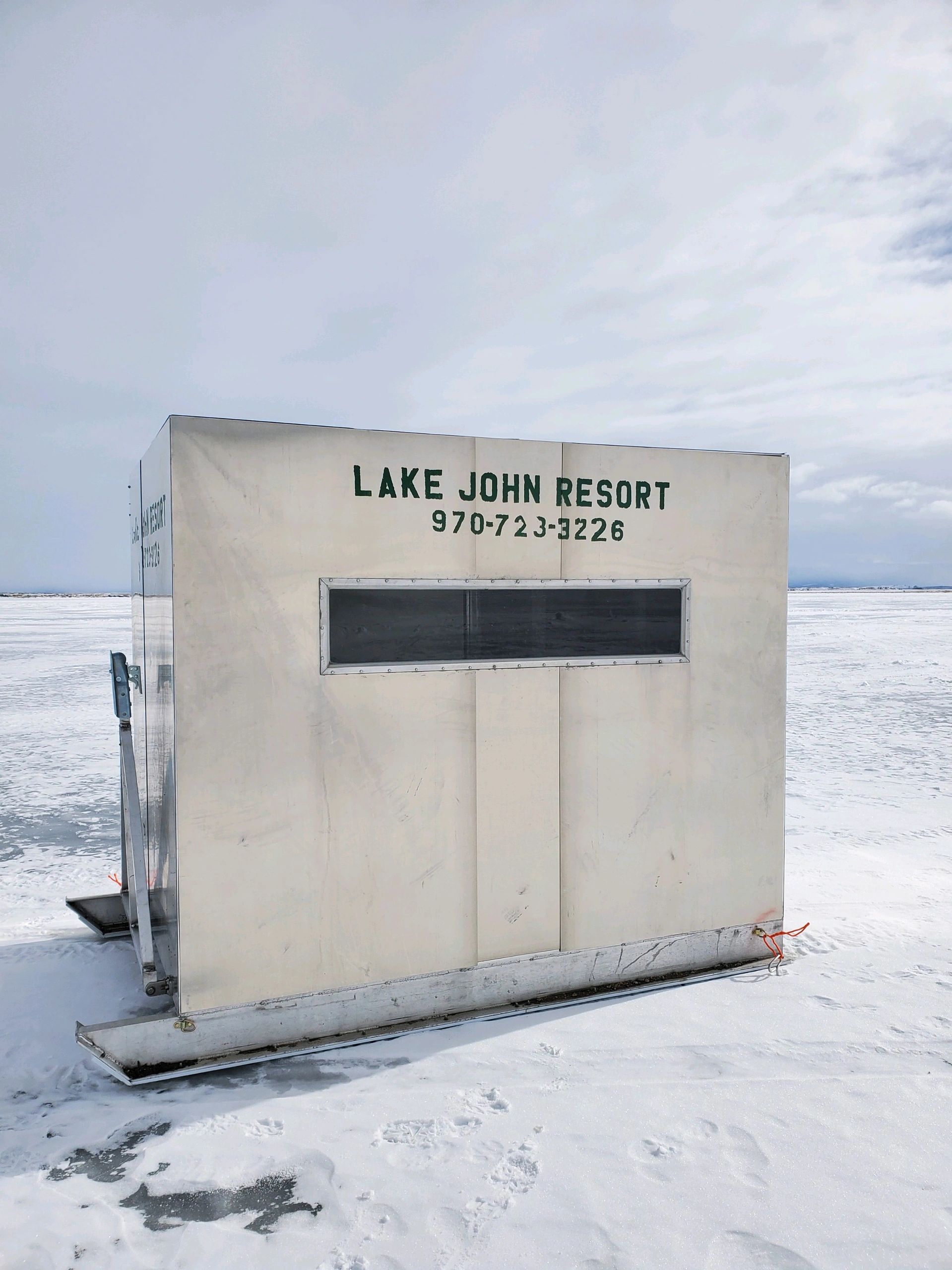 a white ice fishing hut with a window and the words 'Lake John Resort' and a phone number on it