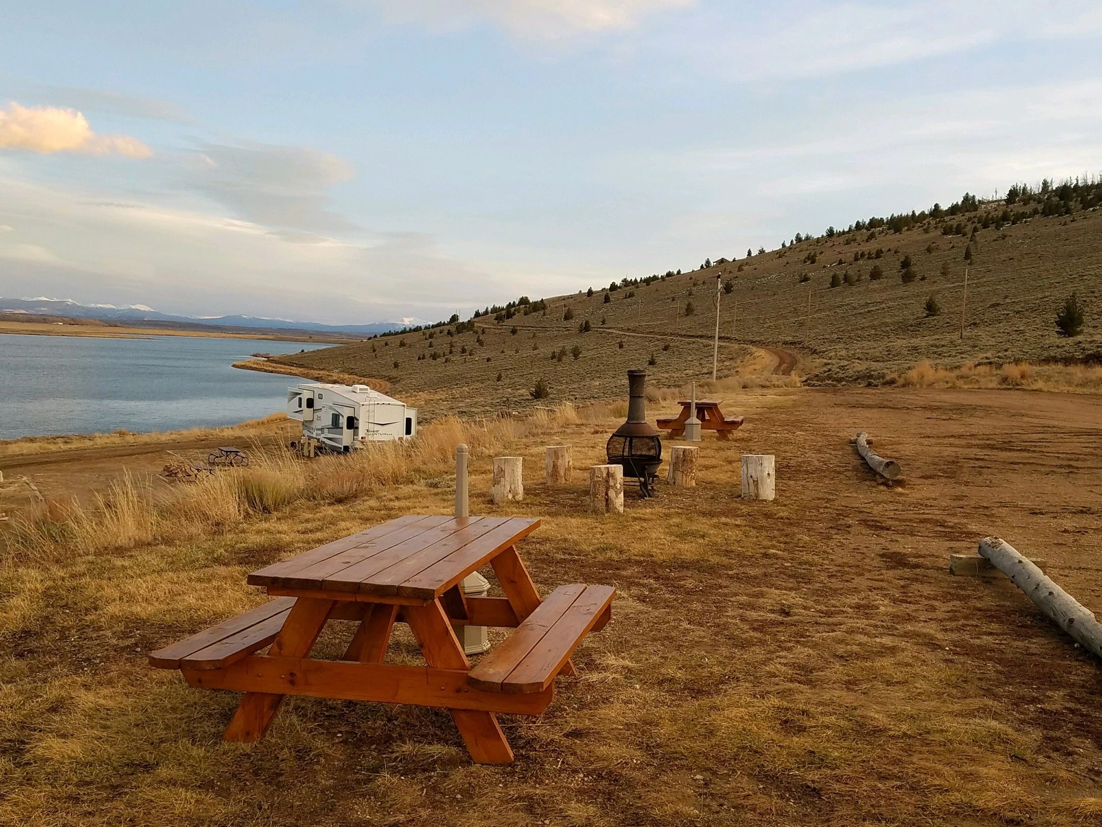 A serene outdoor setting with a picnic table, campfire pit, and a view of a lake and mountains.