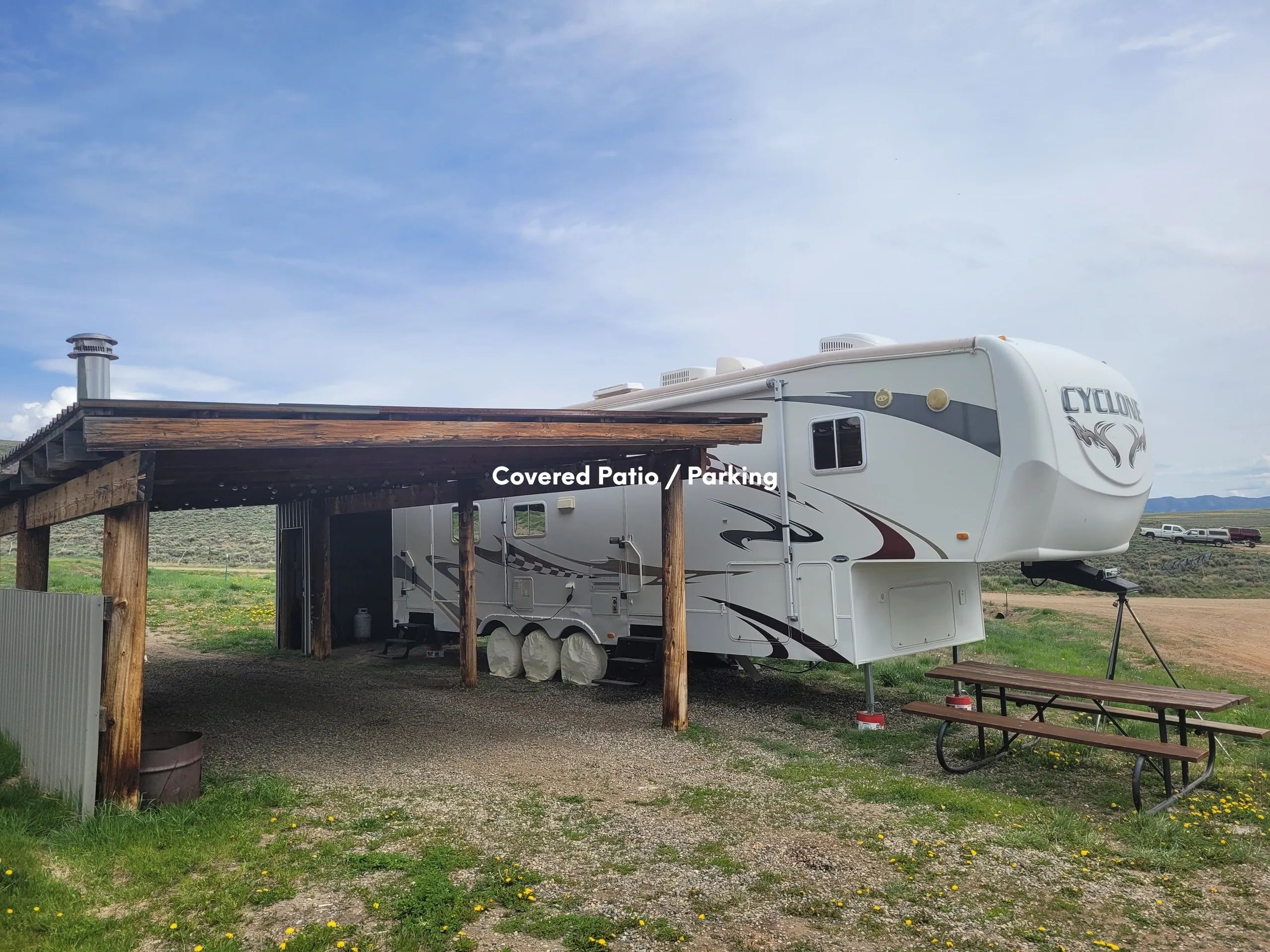 A large white RV parked under a wooden carport with a picnic table in front of it.