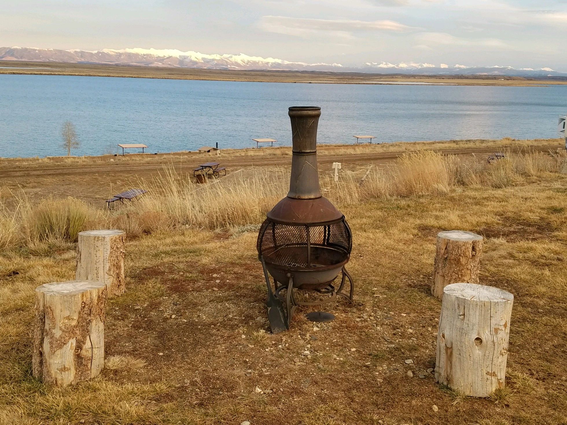 a fire pit with tree stumps around it in front of a lake with mountains in the background