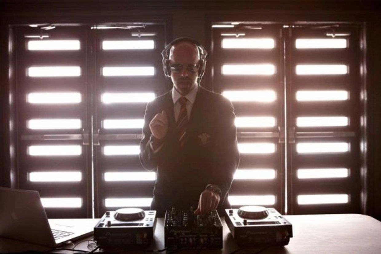 Man DJing in a suit with headphones in front of backlit windows.