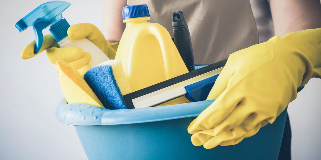 Person holding a blue basket with cleaning supplies wearing yellow gloves.
