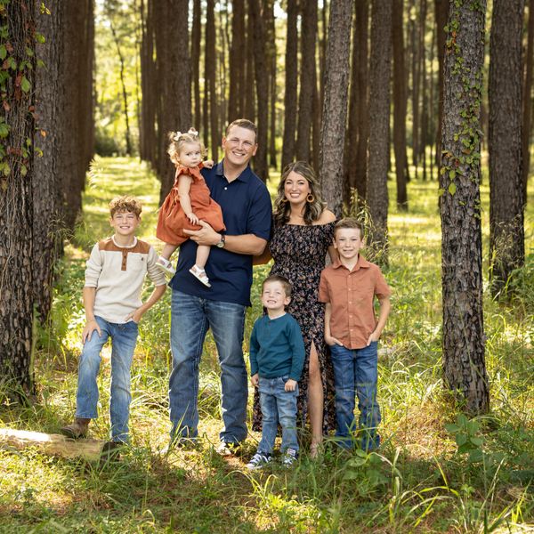 A happy family of six posing in a sunlit forest clearing.