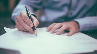 Person writing on documents with a pen on a table.