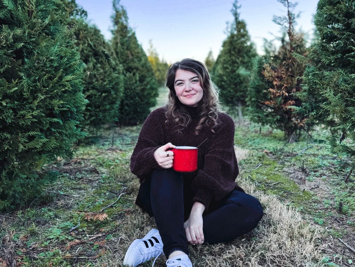 A woman sits among pine trees holding a red mug and smiling.