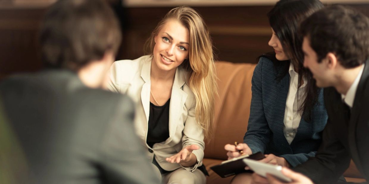 A woman in a white suit speaks to colleagues during a meeting on a couch.