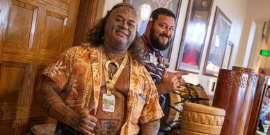 Two men smiling, one with tattoos, posing with wooden drums indoors.