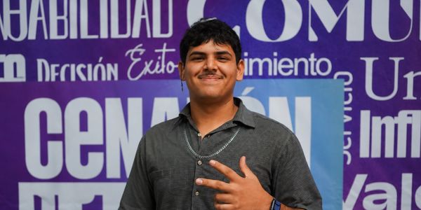 Young man smiling and posing with a peace sign in front of a purple wall with motivational words.