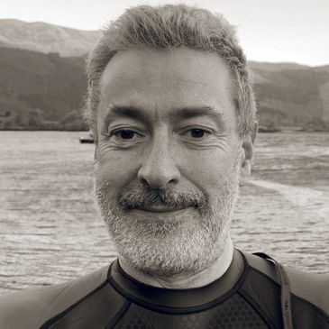 Man with beard smiling near a lake with mountains in the background.