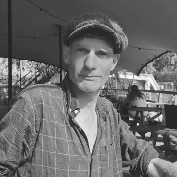 Man in a flat cap sitting outdoors under a canopy.