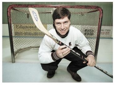 Man posing with a hockey stick in front of a goal on ice rink.