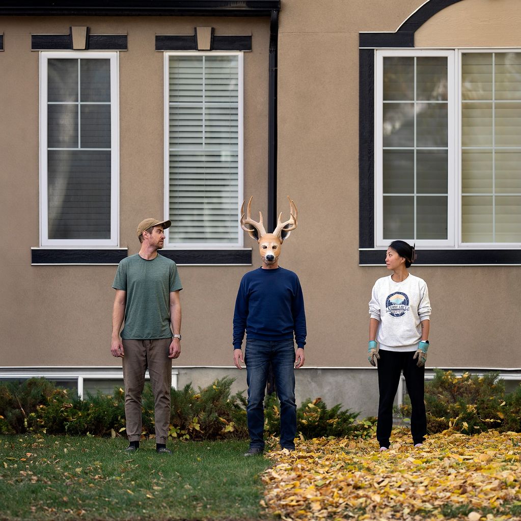 Three people stand outside a house, one wearing a deer mask.