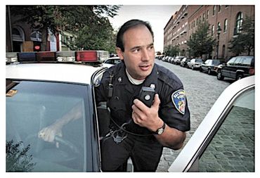 Police officer speaking into a radio beside a patrol car in an urban area.