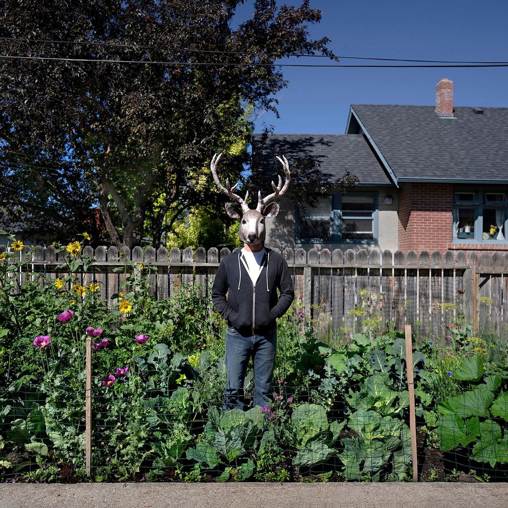 Person wearing a deer mask stands in a garden with flowers and vegetables.