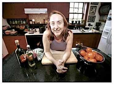 Smiling woman in kitchen with bottles and bowl of tomatoes.