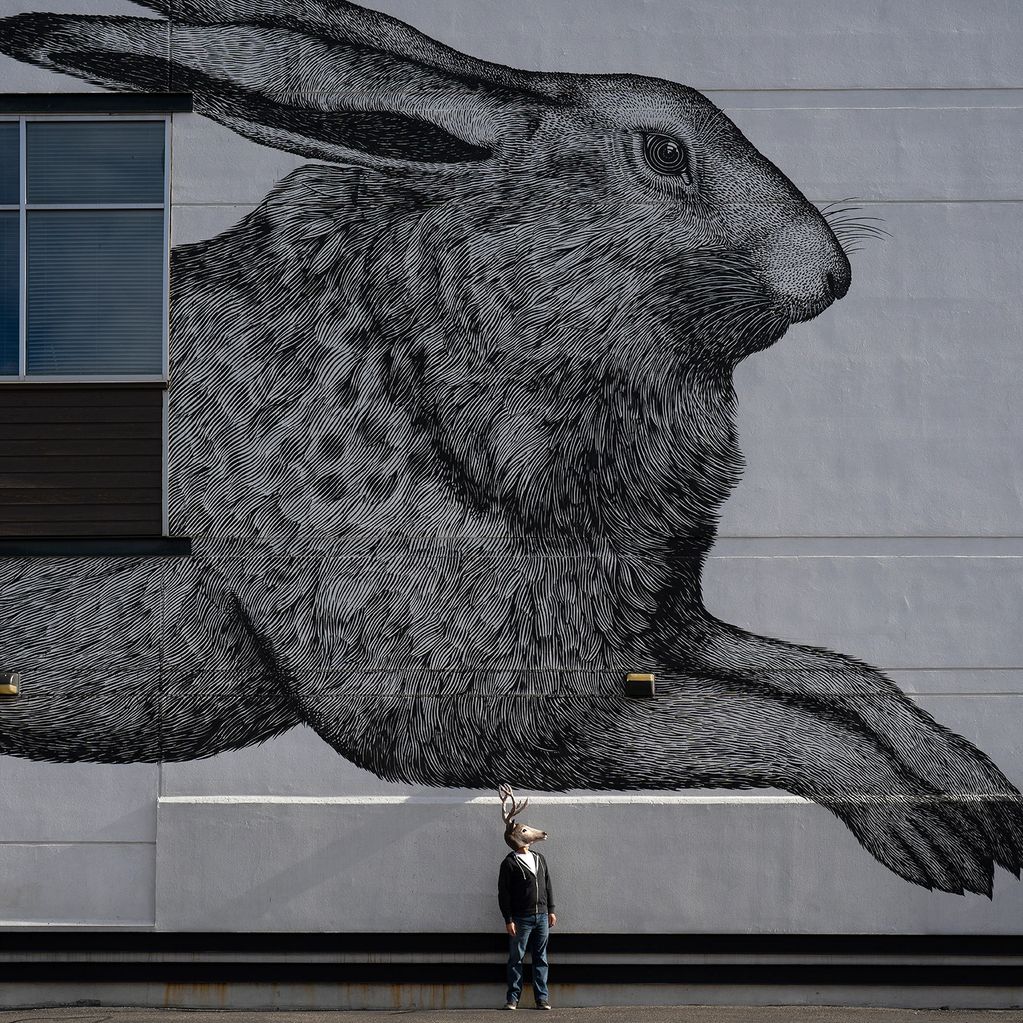 A person with a deer mask stands in front of a giant rabbit mural on a building.