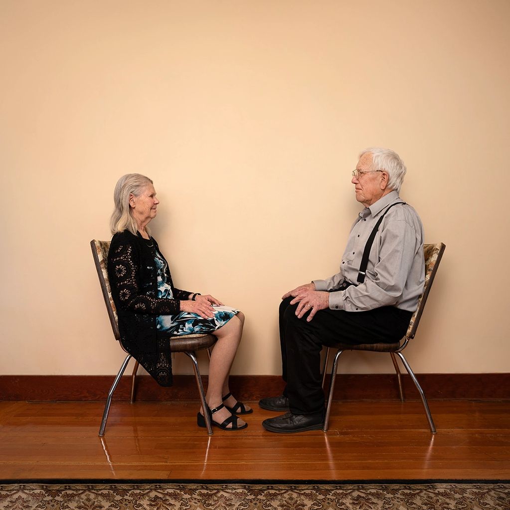 Elderly couple sitting face-to-face on chairs against a plain wall.