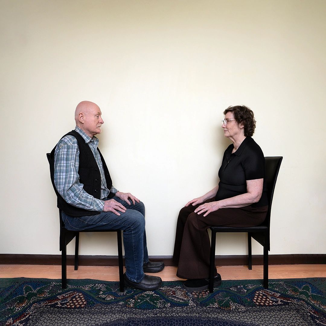 An elderly man and woman sitting face-to-face on chairs in a minimalist room.