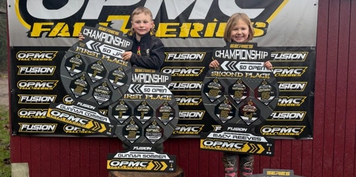 Two kids holding championship trophies in front of an OPMC banner, standing on wooden podiums.