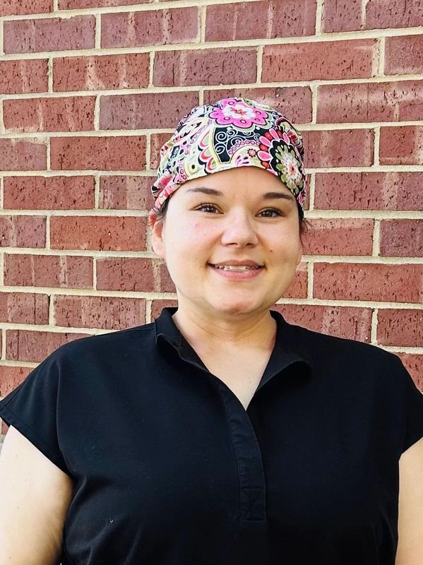 Woman wearing a floral headscarf and black shirt standing against a brick wall.