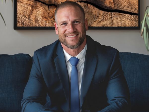 Smiling man in a blue suit sitting on a couch with wooden art behind.