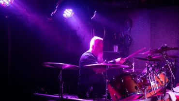 Drummer playing on a stage under purple lights.