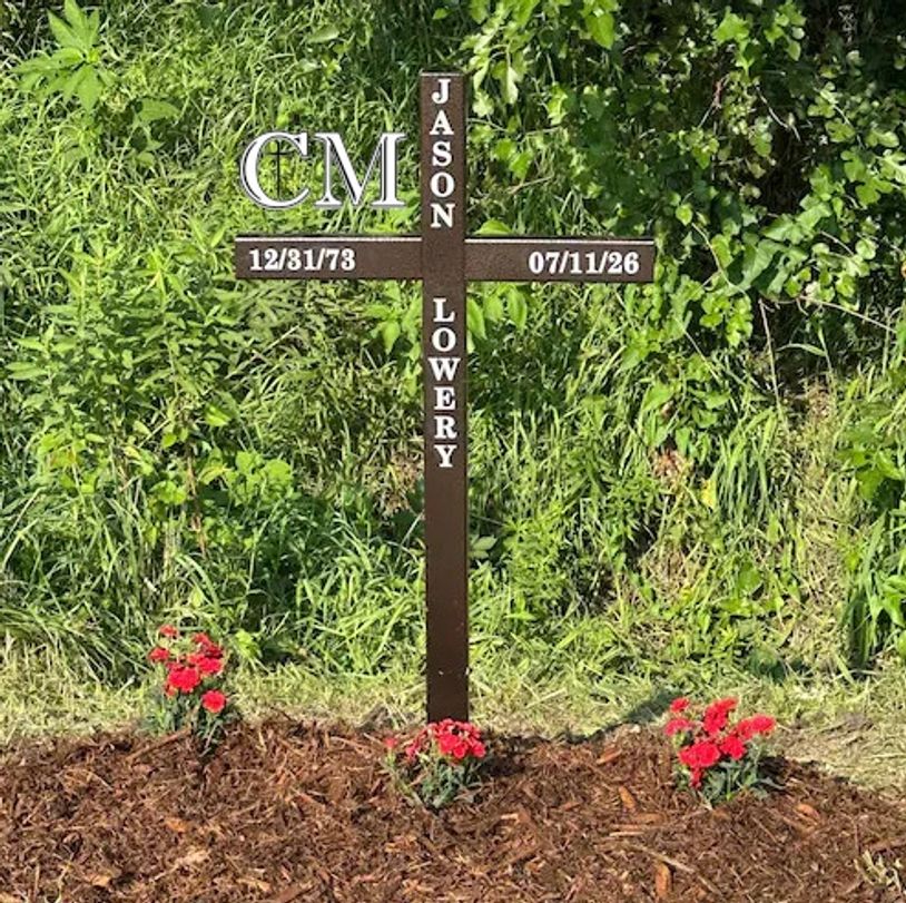 Brown memorial cross for Jason Lowery set in fresh mulch with red flowers in front of green foliage.