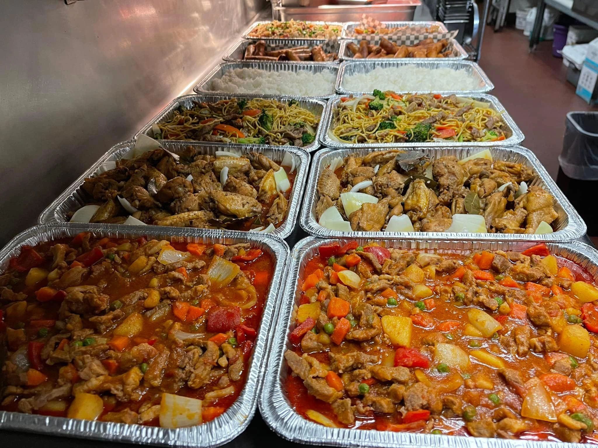 Catering trays filled with various Asian dishes including noodles, rice, chicken wings, and mixed vegetables.