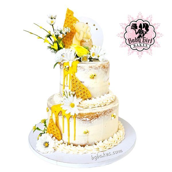 Two-tier honeybee-themed cake with flowers and a bear topper.