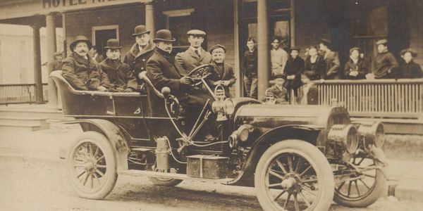 Black and white photograph postcard of an early automobile parked in front of a hotel porch.  The fa