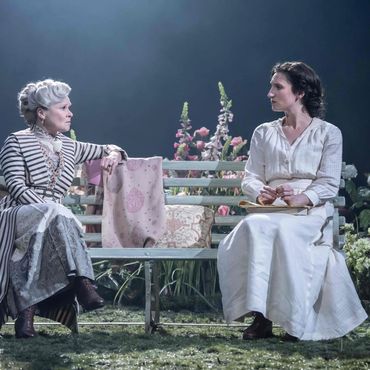 Two women in vintage attire sit on a bench surrounded by flowers under soft lighting.